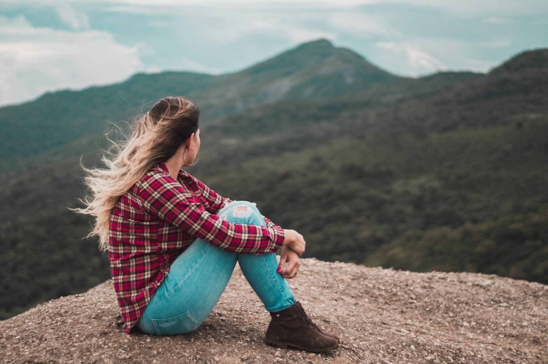 Woman in plaid shirt sitting on a mountain debating what she wants and thinking about how to set realistic goals for her future. 